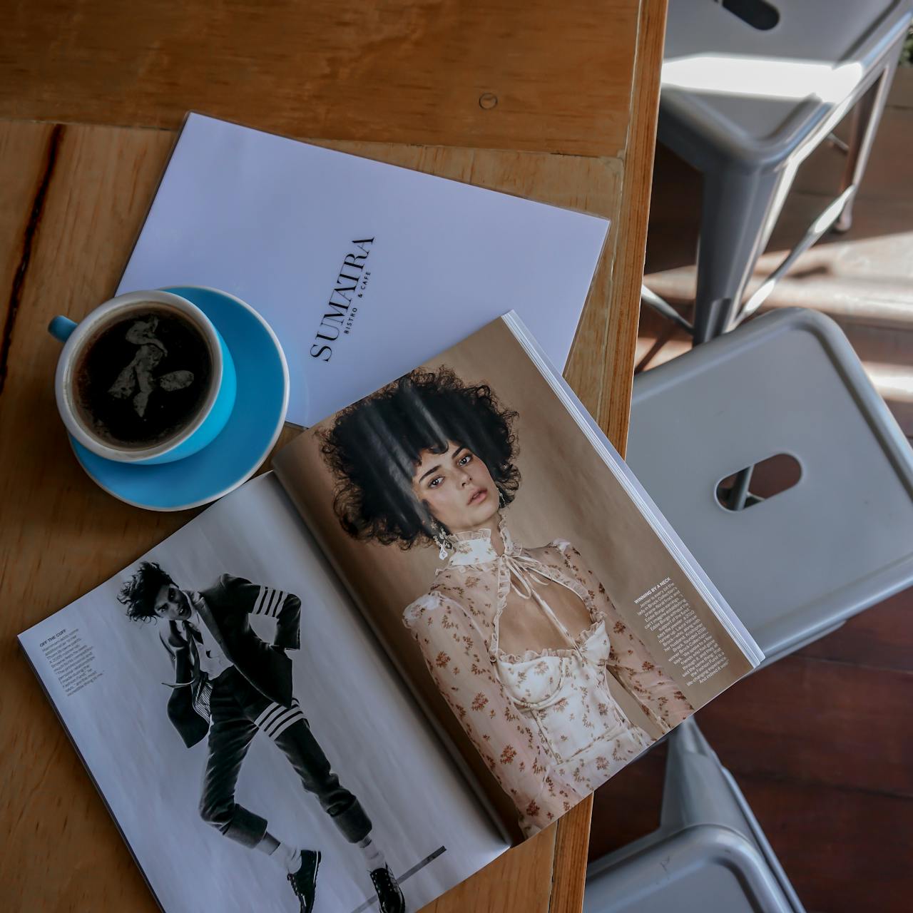 A top view of a blue cup of coffee next to a fashion magazine on a wooden table.